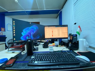 The image depicts a desk setup in what appears to be an office environment. Two computer monitors are prominent, with the left monitor displaying a vibrant blue abstract wallpaper and the right monitor showing a spreadsheet or data interface. In front of the monitors are a black keyboard with Cyrillic and Latin characters, a white Logitech mouse, and a black water bottle. There are also various office supplies such as sticky notes, pushpins, a roll of tape, and a document holder. A corded telephone with a handset is situated to the right of the monitors. A small flag of Tajikistan is visible on the right side of the image, attached to the blinds of a window. The overall lighting suggests an indoor setting, possibly with artificial light sources contributing to the cool blue tone. The scene is a static portrayal of a workspace, with no people actively engaged in an activity. There is no discernible information in the image to indicate the location within the city of Yukhta, Russia.