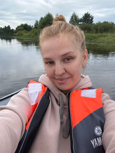 A blonde woman with her hair in a bun is taking a selfie while sitting in a boat on a body of water. She is wearing a light pink hoodie and an orange and grey life jacket with a Yamaha logo. The water is calm with reflections of the cloudy sky and trees on the shore. The background consists of dense green trees and vegetation lining the water's edge. The sky is overcast, suggesting a cool, perhaps humid day. The scene appears to be a natural outdoor environment, possibly a river or lake, within the geographical area of Anteplevo (Okhotinskoe S/P), Russia.