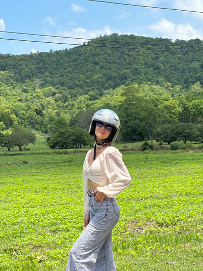 A young woman stands in a bright, green field, a lush, tree-covered mountain range rising behind her under a clear blue sky with scattered clouds. The woman, wearing a white, ruched crop top, light-wash wide-leg jeans, sunglasses, and a silver motorcycle helmet, is positioned in the center of the frame, looking directly at the camera. Her right hand is casually tucked into her jeans pocket. Power lines are visible stretched across the top of the image, above the mountain. The field is densely covered with low-lying green plants, with scattered larger trees in the mid-ground. This scene appears to be in Ham Thuan Bac, Vietnam, a region known for its natural landscapes and agricultural fields.
