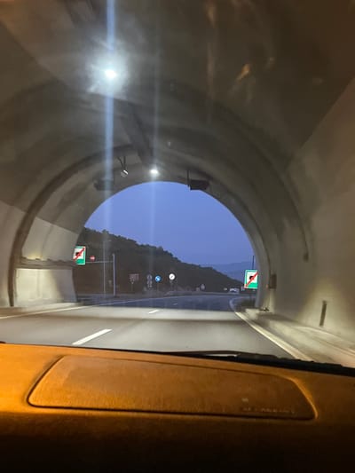 The image shows the interior of a vehicle driving into a tunnel. The tunnel has a concrete arched ceiling with lighting fixtures. On the left side of the tunnel, a green sign with a white square and a red diagonal line is visible, indicating the end of a restriction or a zone. Further down the road, there are other traffic signs and lights. The exterior of the tunnel opens to a hilly landscape under a twilight sky. The dashboard of the vehicle is visible in the foreground, suggesting the perspective is from the driver's seat. The road is paved and marked with white lines. The overall scene depicts a transition from daylight to the darkness of a tunnel.