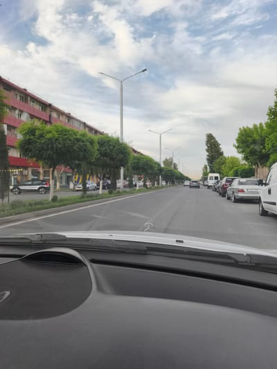 The image depicts a street scene in Artashat, Armenia, viewed from inside a vehicle. A multi-story building with a red facade lines the left side of the street, fronted by a row of neatly trimmed trees. The street itself is asphalt, with a white lane marker. Several cars are parked or driving on the right side of the road, including a white van and a silver sedan with a license plate reading "55 GM 515". Streetlights with multiple lamps are visible along the road. The sky is cloudy with patches of blue. The foreground shows the interior of the vehicle, including the dashboard and a portion of the windshield.