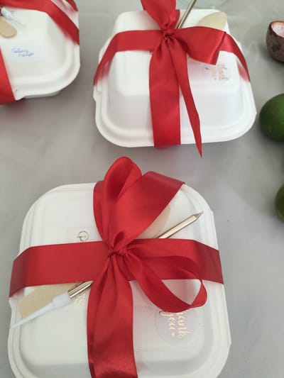 This image features a close-up view of three white, compostable food containers, each adorned with a bright red satin ribbon tied in a bow. Two of the containers are clearly visible, with the third partially obscured in the background. Each container also holds a wooden utensil and a small metallic-colored utensil with a white tip.

On the top left container, a small white label is affixed, containing handwritten text that appears to be "Főztük nagy". On the middle container, a circular sticker with the text "Thank you" in a cursive font is visible. A similar "Thank you" sticker is present on the bottom container, though it is partially covered by the ribbon.

The setting appears to be an indoor event or gathering, likely a catered meal or a gift presentation. The neutral background suggests a table setting. The presence of limes and what looks like the rind of a passion fruit in the background might hint at the type of food or beverages being served. The lighting is soft and even, suggesting an indoor environment, possibly during the day. There are no people visible in the frame, focusing the attention solely on the beautifully presented food containers. The overall impression is one of thoughtful preparation and a welcoming gesture.