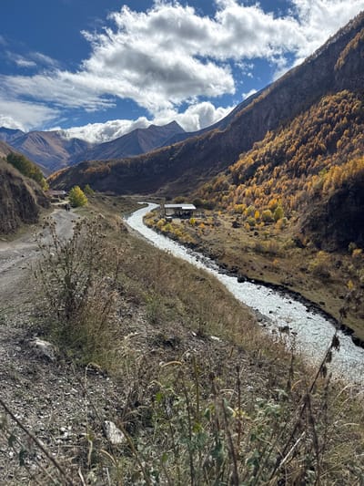 The image displays a mountain valley scene located in the city of Kvemo Okrokana, Georgia, under a blue sky with scattered white cumulus clouds. A dirt road occupies the left foreground, ascending into the distance and featuring loose gravel and dry, sparse vegetation. A fast-flowing, light-colored river traverses the valley from the upper left, curving towards the lower right. Along the river's right bank, a structure with a dark roof and an attached covered area is visible. Further up the valley, on the left side of the dirt road, a second building with a red roof is partially obscured. Two human figures are discernible on the dirt road, walking away from the camera, positioned near the red-roofed building. The mountain slopes flanking the valley are densely covered with deciduous trees displaying vibrant golden-yellow and orange autumn foliage. The background consists of rugged mountain ranges, with some peaks partially concealed by clouds. The immediate foreground features dry grasses and tall, spiky plant stalks.