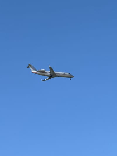 The image displays a light-colored passenger airplane captured against a clear, bright blue sky. The aircraft is angled from the bottom left towards the top right of the frame, suggesting it is either ascending, descending, or making a turn. Its two engines are mounted on the rear fuselage, typical of a regional jet.
A notable detail is that the landing gear appears to be deployed, visible underneath the fuselage and wings, which indicates the plane is in the process of taking off or landing. The aircraft's body is predominantly white or light gray, with a distinctive red stripe visible along the lower part of its tail section. The sky is uniformly clear and cloudless, signifying good weather conditions during the daytime, likely mid-day to afternoon.
The perspective is from below, looking upwards at the plane. No other objects or visible text are present in the sky or on the aircraft. The location context provided is Middelkerke, Belgium.