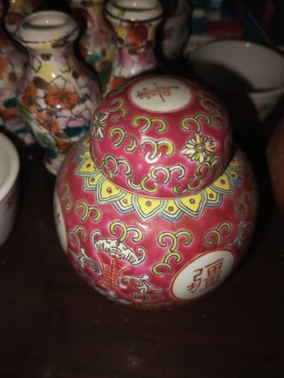 A red, round Chinese ginger jar with a lid sits on a dark surface. The lid features flowers and swirling patterns in yellow and green. The jar itself has similar designs with yellow and light blue accents. Chinese characters are painted in white circles on the jar's surface. Additional miniature, colorful vases are present in the background along with a white teacup.