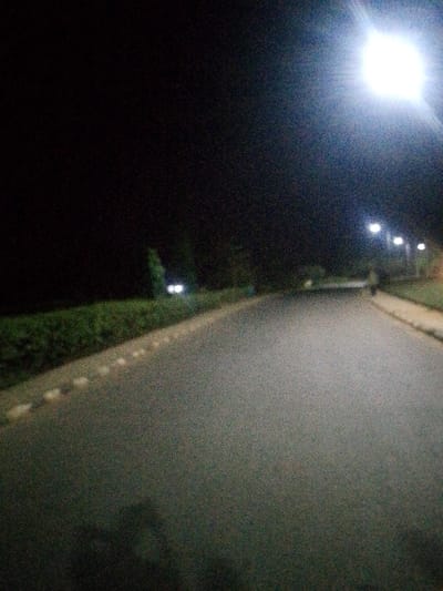 A dark road stretches into the distance under the dim light of street lamps. A bright light, possibly from a lamp, dominates the upper right corner. Hedges and a curb line the left side of the road, while a walkway runs along the right. A pair of lights is visible further down the road, perhaps from another vehicle. The scene is dark and quiet, illuminated only by artificial light sources.