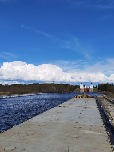 A view of the Svir River in Анхимово, Russia, presents from the stern of a barge, displaying a gray, weathered deck with numerous hatches. The dark-blue water of the Svir flows to the left, bordered by a long, concrete structure. Beyond, a forested horizon is broken by an electricity pylon. Across the river, a canal leads to a pair of lock towers built in white with red doors. The sky is a vibrant blue, populated with scattered cumulus clouds.