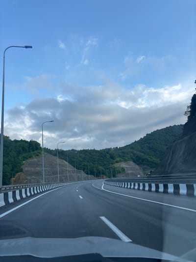 The image shows a view from a vehicle traveling on a highway in Surami, Georgia. The highway curves to the right, bordered by a concrete barrier with alternating black and white stripes. On the left side of the highway, a steep, tree-covered mountainside rises, with terraced sections and streetlights visible. On the right side, the mountain slopes more dramatically, with exposed rock and concrete reinforcement. The sky above is partly cloudy with patches of blue sky visible. The lighting suggests it is either early morning or late afternoon. No people or other vehicles are clearly discernible in the immediate foreground or on the highway.
