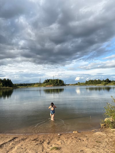 The image depicts a picturesque natural landscape with a calm body of water, likely a lake or a large pond, surrounded by lush green trees. A sandy shore is visible in the foreground. A young person, wearing a dark blue swimsuit with a floral pattern, is standing in the shallow water, facing away from the camera. They appear to be holding a mobile phone or camera in their hands, possibly taking a photograph or video. The sky is dramatic, filled with a mix of dark, heavy clouds and patches of blue sky, suggesting an overcast or impending weather change. In the background, on the far bank of the water, a dense forest stretches across the horizon, with several tall, industrial-looking towers visible above the treeline. The overall scene conveys a sense of tranquility and natural beauty, interrupted by the presence of man-made structures. The location is identified as being in the city of Zamost'e, Russia.