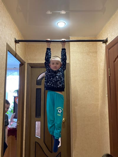 A boy is hanging from a metal bar installed in a doorway. He is wearing a dark patterned sweatshirt and green sweatpants. Another person is visible through the open doorway below, partially obscured by objects, but no other information about the location in Bely Klyuch, Russia, can be ascertained from the image.