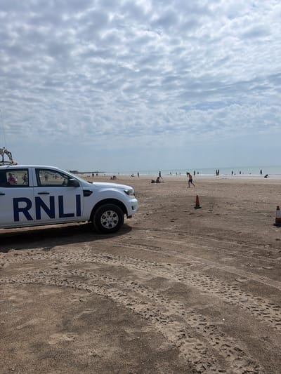 The image depicts a white RNLI (Royal National Lifeboat Institution) vehicle parked on a sandy beach. The vehicle has large blue lettering on its side that reads "RNLI". The scene is set on a beach with the sea visible in the background. There are several people on the beach, some walking near the water's edge and others scattered further back. The sky is partly cloudy, suggesting a mix of sun and clouds. The ground is covered in sand with visible tire tracks and footprints, indicating recent vehicle and human activity. There are also orange traffic cones on the sand. The overall impression is of a busy beach on a day with fair weather. The perspective of the image is from a low angle, looking up towards the sky and across the beach. The time of day appears to be daytime, likely afternoon, given the lighting. The location cues (RNLI vehicle, beach, sea) point to a coastal setting.