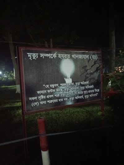The image shows a dark, outdoor scene at night, likely in Bagerhat, Bangladesh, given the context. A signboard is prominently displayed, featuring white text on a dark background. The text appears to be in Bengali and includes the phrase "মৃত্যু সম্পর্কে হযরত খানজাহান (রহ)" which translates to "About Death of Hazrat Khan Jahan (R.)". Below this title, there is a poem or quote with several lines. The signboard has a lit candle graphic above the text. The signboard is framed by a dark border and supported by a dark structure, partially visible. In the foreground, a section of a red and white striped pole is visible, along with a thin wire or rope stretched horizontally across the frame. The background is dark and blurry, with hints of trees and foliage. The overall impression is of a memorial or informational display set up in a park-like environment.