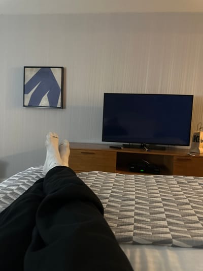 A person lies on a bed with patterned gray and white bedding, wearing black pants and white socks. A wall-mounted television sits above a wooden cabinet, while a framed abstract artwork hangs nearby. The wall is a light gray with subtle horizontal lines.