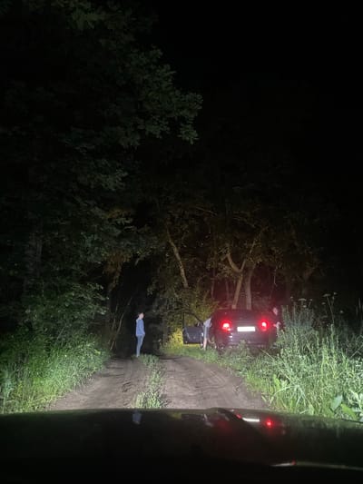 A night-time, low-angle, wide shot captures a dark, dirt road in a wooded area, possibly on the outskirts of Yekaterinovka, Russia. The scene is illuminated by headlights from the viewer's perspective and by the car's taillights. A dark-colored car is parked on the right side of the road with its driver's side door open. Two individuals are near the car, one standing to the left and appearing to be getting into or out of the vehicle, and another to the right, partially obscured by foliage, possibly attending to the car. A third person stands further down the road to the left, facing away from the camera and towards the car. Tall, overgrown grass and trees flank the road, creating a dense, dark, and somewhat isolated atmosphere. The road surface appears wet and muddy in places, indicating recent precipitation or damp conditions.