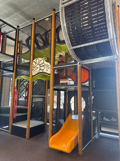 The image captures the interior of a children's indoor playground, possibly located in Placerville, United States of America, as evidenced by the style of play structure common in fast-food restaurants. The playground features a multi-level structure constructed with metal mesh, wooden beams, and colorful plastic elements. A young boy wearing a red shirt and green shorts is visible within the structure, appearing to be climbing or playing. An orange slide curves down from an upper level to the carpeted floor. There are stairs with red handrails, a green-colored net-like obstacle, and a warning sign indicating "No climbing on outside of the playground equipment." In the background, shoes are placed on a shelf. The overall environment suggests a recreational area designed for children's activities.