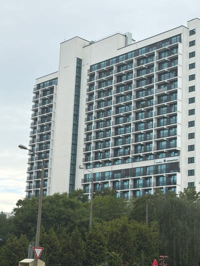 The image shows the modern facade of a large white apartment building in Halle (Saale), Germany. The building is tall and features numerous balconies with black railings, each adorned with blue-tinted windows. The architecture is geometric and repetitive, with a grid-like pattern of windows and balconies. In the foreground, lush green trees and foliage partially obscure the lower levels of the building. Two streetlights are visible on either side of the building. On the lower right side of the building, a sign reads "Sparkasse," a common German savings bank. The sky is overcast, suggesting a cloudy day.