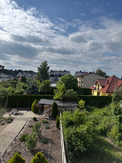 This image depicts a suburban backyard scene in Marki, Poland. The setting is a residential area with multiple houses and lush greenery. The foreground features a paved walkway and a garden area with mulch, small shrubs, and some larger manicured bushes. A small, rustic shed or outbuilding is visible in the middle ground.
Beyond the immediate garden, several houses are partially visible. One house, painted yellow with a red tile roof, is prominent in the upper portion of the frame. Other houses with various rooflines and architectural styles are seen further back, interspersed with dense trees and hedges, suggesting a well-established neighborhood.
The sky above is a mix of blue and white clouds, indicating daytime with partly cloudy weather. The sunlight appears to be casting shadows, suggesting it could be late afternoon or early morning. There is no activity or event taking place; it's a static view of a backyard. No people are visible, and no text is present in the image. The overall impression is one of a peaceful, green, and well-maintained residential environment.