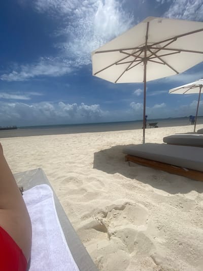 View from a lounge chair at the beach in Marina Maroma, Mexico. A white umbrella is visible to the left, and blue sky and clouds are in the background. The beach is white sand with some lounge chairs on it. The ocean water is visible in the distance. The legs and red bathing suit of the person lying in the lounge chair are visible in the foreground.