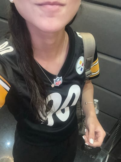An individual is depicted from the upper torso to the lower face in an indoor setting within the city of Santa Catarina, Mexico. The person wears a black Pittsburgh Steelers NFL jersey with yellow shoulder stripes and the number "28" displayed in white on the front. A silver chain necklace adorns the neck, featuring an NFL shield pendant and a small pink heart charm. Small white pearl earrings are present. A beige backpack strap is visible over the right shoulder. The right hand is partially visible, showing white nail polish on the fingernails and a silver-colored bracelet on the wrist. In the background, dark grey vertical panels form the wall, and a reflective dark floor is partially visible. A clear plastic water bottle stands in the lower right background. No specific actions or interactions are discernible beyond the individual being present and partially visible.