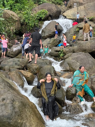 The image depicts a rocky waterfall area in Jaflong, Bangladesh, with several people enjoying the natural surroundings. In the foreground, two women are seated on wet rocks near the cascading water. The woman on the left wears a black outfit and a patterned scarf, with her feet in the water. The woman on the right is dressed in a turquoise and gold shalwar kameez and is also partially submerged.

Further up the rocks, a group of people are visible. A man in a black t-shirt and shorts stands on a rock, facing away from the camera. Another man in a yellow and black checkered shirt is also present. A young boy in a yellow t-shirt and shorts is standing on a rock near the waterfall. Other individuals are scattered across the rocky terrain, some appearing to be taking photos or observing the waterfall. The environment is characterized by large, moss-covered boulders and lush green vegetation growing on the slopes. The water is clear and flowing over the rocks, creating a dynamic and natural setting. The overall atmosphere suggests a recreational outing to a scenic spot.