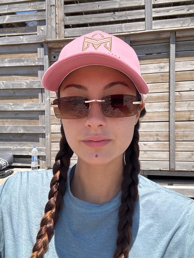 In the city of Moudros Municipal Unit, Greece, a young woman with dark hair braided into two pigtails is captured in a close-up portrait. She wears a pink baseball cap with a gold emblem and stylish rimless sunglasses. Her face is tilted slightly upwards, and her lips are slightly parted. A mole is visible on her chin. She is dressed in a light blue, long-sleeved shirt. The background consists of a wooden slatted structure, possibly part of a beach or outdoor setting. To the left of the woman, a plastic bottle of water stands on a flat surface. The lighting suggests it is daytime, likely sunny.