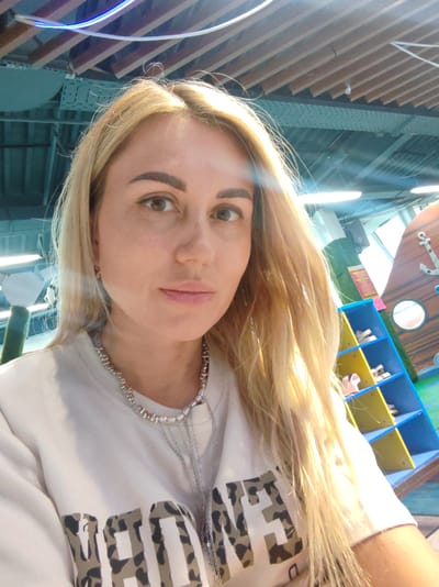 A young woman with long, blonde hair and light skin is taking a selfie. She is wearing a white sweatshirt with a leopard print design on it. A silver necklace with multiple charms is visible around her neck. She has light brown eyes and appears to be smiling slightly. The background shows a brightly lit indoor space with wooden ceiling beams and fluorescent lights. To the right, there is a colorful display shelf with shoes on it, and a wooden structure resembling a ship's hull with an anchor. The overall setting suggests a children's play area or an amusement park. The lighting indicates it is daytime. There is no visible text in the image.