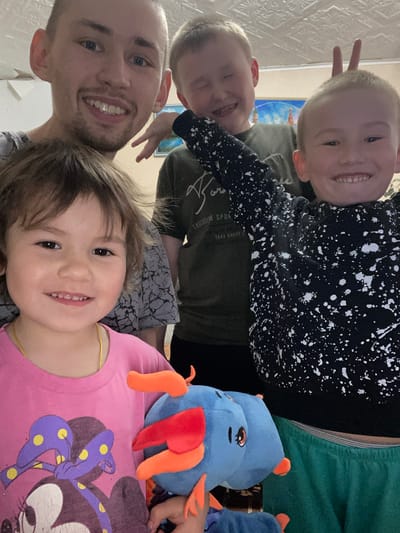 A selfie shows a fair-skinned man with light hair and a beard, smiling at the bottom left. Above him are three children with fair skin and light hair, also smiling. One child holds up two fingers behind another child's head. A stuffed blue animal with orange and red accents lies next to the child in the pink shirt. The environment shows a room with wallpaper, and the scene appears to be indoors, possibly in Bely Klyuch, Russia.