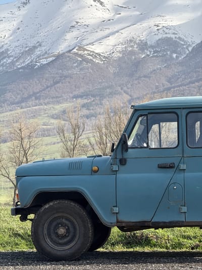 The image captures a light blue vehicle, likely a truck, parked alongside a road in Margahovit, Armenia. The tire of the vehicle is visible, set against a backdrop of green grass and an asphalt road. In the distance, a mountainous landscape is visible, partially covered in snow, juxtaposed against valleys and sparse trees, indicating a rural or mountainous setting.
