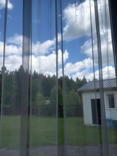 The image presents a view through sheer curtains, looking out onto a rural or suburban landscape. The foreground is dominated by the vertical lines of the translucent gray curtains, which obscure portions of the scene. Beyond the curtains, a bright blue sky with scattered white cumulus clouds fills the upper portion of the image. Below the sky is a dense line of dark green trees, forming a forest or thick woodland. To the right of the trees, a small, light-colored building with a gray, corrugated metal roof is visible. The building has a dark-colored door and a window with a white frame. In front of the building, a small blue table and chair are visible on a green grassy lawn. The lawn extends to the foreground, appearing somewhat unkempt with scattered small white flowers. The scene suggests a daytime view, likely during fair weather. The location in Podzhigorodovo, Russia is not directly evident from visual cues within the image itself, but is provided as contextual information. There are no people or animals visible in the image.