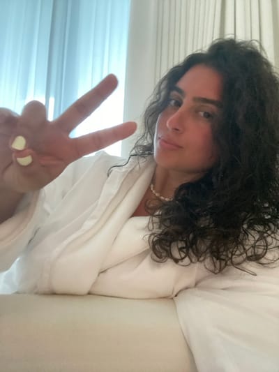 A person is wearing a white robe and a pearl necklace, making a peace sign with their left hand. The location is not El Caracol, Mexico.