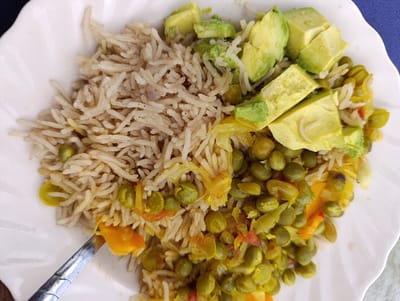 An overhead, close-up shot captures a wholesome meal served on a white, fluted-edge plate. The plate is filled with a variety of ingredients, prominently featuring light brown, long-grain rice, which occupies the upper-left half of the plate.
Adjacent to and mixed with the rice, and forming a larger portion on the bottom and right, are numerous cooked green peas or pigeon peas. These peas are interspersed with other cooked vegetables, including soft, translucent yellow pieces, likely sautéed onions or cabbage, and bright orange chunks that resemble carrots or pumpkin.
Crowning the upper-right section of the dish are several freshly diced pieces of light green avocado, adding a vibrant contrast and creamy texture. A silver spoon is visible in the lower-left corner, partially submerged in the food, suggesting the meal is either about to be eaten or is already in progress.
No people are visible in the frame, and there is no discernible text. The setting appears to be a casual dining arrangement. The time of day and weather cannot be determined from the image. The combination of rice, peas/beans, and avocado is consistent with local cuisine in Kaplelwo, Kenya, the provided location context.