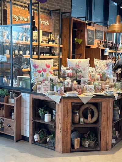 This image displays the interior of a retail establishment in Altenheim, Germany, characterized by a blend of rustic and modern design. The central focus is a large, rectangular wooden display table, constructed from stacked crate-like structures, positioned on a light-colored, smooth floor.
The top surface of this table is covered with a light fabric runner and densely arranged with merchandise. Prominent items include three square throw pillows featuring vibrant floral patterns (tulips and mixed wildflowers), numerous glass candle holders, votives, and small vases, many adorned with floral designs. Various small packaged gift items and a vertical sign with the German text "Du musst dein Anderes Leben" are also present.
In the background, against a stone wall, are dark metal shelving units with wooden shelves. To the left, a glass partition reflects outdoor elements and partially obscures more shelves holding dark-bottled products, with a small white ceramic chicken figurine visible behind it. To the right, wooden signs reading "regional" and "frisch" (fresh) are affixed to the shelving units, which display a variety of bottled goods including a large bottle, champagne, and numerous smaller bottles. Further right, more dark wooden shelves feature chalkboards labeled "Küche" (Kitchen), "Schönes" (Beautiful), and "Getränke" (Drinks), showcasing additional bottled items. A copper-toned pendant lamp hangs from the ceiling in the upper right.
The lower sections of the central display table, configured as open shelves, contain decorative arrangements. These include potted green plants, a white lidded container, a large glass jar filled with granular material, a natural fiber wreath with a small wooden utensil, two white pillar candles, and various baskets holding greenery and a wooden brush. No individuals are present, and no actions or interactions are occurring.