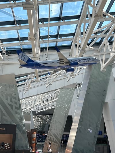 The image depicts the interior of a modern airport terminal, characterized by its expansive glass roof and complex white metal support structure. Suspended from the ceiling is a model airplane, prominently displaying the "Belavia" logo and registration number "EW-455PA" on its tail. The airplane model is a central focal point, positioned against the backdrop of the architectural elements of the terminal.
In the foreground and midground, large, angled glass panels with abstract patterns are visible, some appearing to be decorative or structural elements of the terminal's design. These panels create a sense of depth and reflect the surrounding environment.
On the lower levels, glimpses of shops or service areas are visible, with signs and displays indicating commercial activity. People are present within the terminal, though their presence is secondary to the architectural and model airplane features. One individual is seen walking in the lower right portion of the image, and others are faintly discernible in the background, suggesting the ongoing operations of an airport.
The overall impression is one of contemporary airport architecture, showcasing both functional design and decorative elements. The specific location within the city of Пекалінскі сельскі Савет, Belarus, is not explicitly indicated by any visual cues in the image itself, but the presence of the Belavia airline suggests this is likely Minsk National Airport.