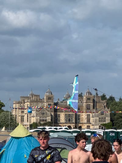 A vibrant festival scene unfolds beneath a cloudy sky. A stately stone building adorned with colorful banners provides a backdrop. Tents dot the foreground, partially obscuring several shirtless young people. A distinctive flag featuring a blue and white cross flutters in the breeze above the crowd. The atmosphere suggests a lively outdoor event.