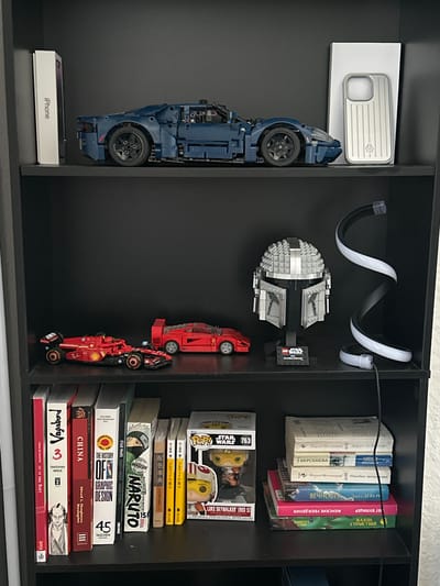 The image displays a dark bookshelf filled with various items. The top shelf features a blue Lego car model and an iPhone box. The middle shelf holds two smaller red Lego car models, a Lego Star Wars Mandalorian helmet, and a spiral LED lamp. The bottom shelf is crowded with books, including titles in English, Japanese, and Russian, as well as a few collectible figurines. The lighting is dim, and the overall setting appears to be a domestic environment. The image does not contain any specific information or visual cues that would allow for the identification of the city of Bielefeld, Germany.