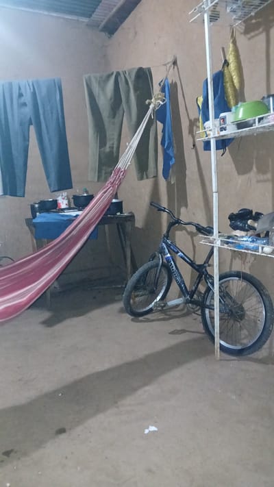 The image depicts an indoor scene in El Samán, Venezuela. A bicycle is leaning against a white metal drying rack. Underneath the rack are items including clothing and a green container. A hammock, striped in red and white, is slung across the room. Other articles of clothing appear to be hanging. A stove with cookware on top is also visible. The floor is a uniform tan color.
