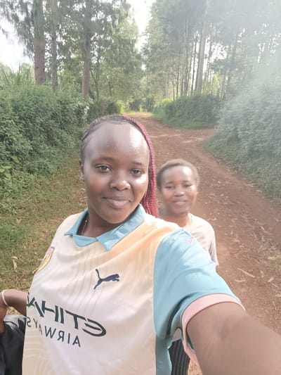 The image features two women outdoors in Kaplelwo, Kenya, appearing to take a selfie. The woman in the foreground is centered and looking directly at the camera with a gentle smile. She has dark skin and reddish-brown braided hair pulled back from her face. She is wearing a light-colored (pale yellow/cream and light blue) athletic jersey. Visible text on her jersey includes "ETIHAD AIRWAYS" in black capital letters, with "WHITE" slightly obscured, and a black Puma logo. Her right arm is visible in the lower right corner, indicating she is holding the camera.
Behind her, partially visible and slightly out of focus, is another woman also looking towards the camera with a subtle smile. She is wearing a plain light-colored t-shirt.
The setting is a natural, outdoor environment. They are standing on a dirt road or path that recedes into the background. The sides of the path are lined with dense, green bushes and numerous tall trees, suggesting a rural or wooded area. The lighting is soft and diffused, indicating it is daytime, possibly under an overcast sky or during the softer light of morning or late afternoon. The overall mood is casual and pleasant.