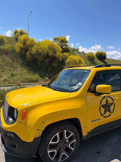 A yellow Jeep Renegade is parked on an asphalt surface. The vehicle has a large black star decal on the rear passenger door and "RENEGADE" badging. The front fender has visible bolts and is slightly detached. The background features a grassy hillside covered in blooming yellow shrubs. A lamppost is visible on the hillside, and a clear blue sky with scattered white clouds stretches above. The scene is illuminated by bright sunlight. The location is identified as Sagholasheni, Georgia.