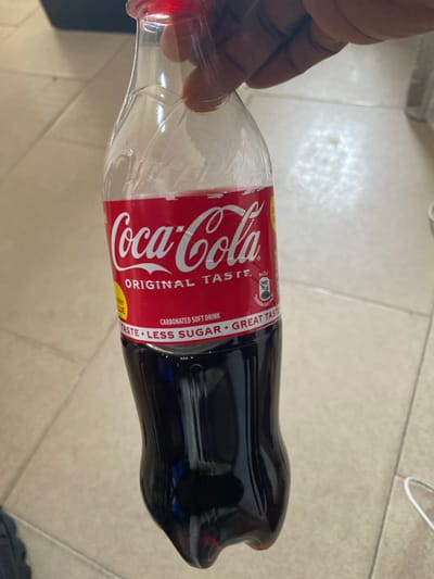 A dark-skinned hand grips the neck of a clear plastic Coca-Cola bottle, which contains a dark brown carbonated soft drink. The bottle features a red label with white "Coca-Cola ORIGINAL TASTE" branding and text indicating "LESS SUGAR." The bottle is approximately two-thirds full and has a red plastic cap. The scene is illuminated, with reflections visible on the bottle and the light-colored tiled floor in the background. The environment is an interior space in the city of Akarakumoh, Nigeria, characterized by light-colored floor tiles and visible grout lines, with a white cable partially discernible on the far right.