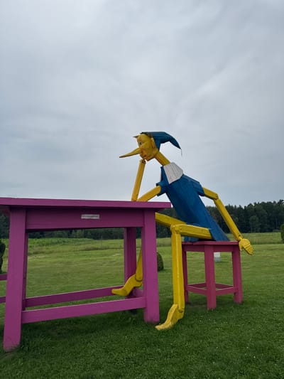 In Skapiškiai, Lithuania, a large wooden sculpture of a whimsical figure, resembling Pinocchio, sits at a pink table and stool. The figure has a yellow body with blue clothing and a blue hat. Its long nose and facial features are prominent. The figure is posed as if deep in thought, with one arm resting on the table. The table and stool are bright pink and are set outdoors in a grassy field. In the background, there are trees and a cloudy sky. A sign on the table reads "LIPTI DRAUDŽIAMA," which translates to "Climbing is forbidden."