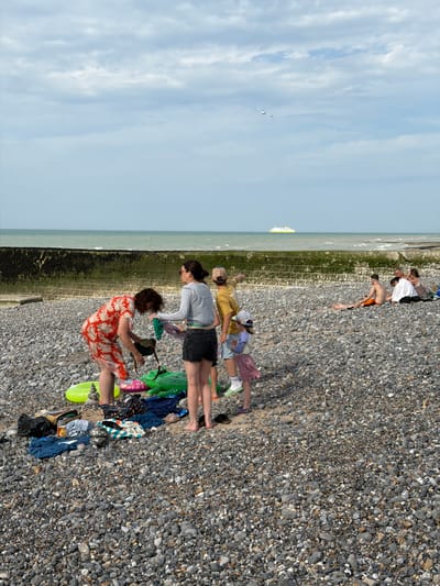 The image depicts a group of people enjoying a sunny day at a pebble beach in Hautot-sur-Mer, France. The scene is oriented vertically, with the sky taking up the left portion of the frame and the beach filling the rest. Several individuals are scattered across the pebbles, some reclining and others interacting with each other.

In the foreground, two women are visible. One is wearing a red and white patterned dress and appears to be tending to something on the ground, possibly a dog or a bag. The other woman, in a grey long-sleeved shirt and dark shorts, is seated beside her. Further up the beach, a young boy in orange shorts is lying down, and behind him, a few other individuals are partially visible, including one wearing a white shirt.

Scattered amongst the people are various beach items, including towels, bags, and inflatable toys. The sea is visible in the background, with a ferry or boat on the horizon. The weather appears to be clear and sunny, indicated by the bright sky and the casual attire of the people. The overall mood seems relaxed and leisurely, consistent with a day at the beach. No visible text is present in the image.