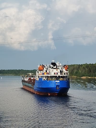 A blue and red cargo ship labeled "PYHA" is navigating a waterway, likely a river or canal, near Gari, Russia. The ship is moving away from the viewer. Its deck is white with various structures and orange life rafts. Dense green forest lines the shores. The sky is partly cloudy with visible power lines extending horizontally across the upper part of the frame. The water is dark with visible ripples.