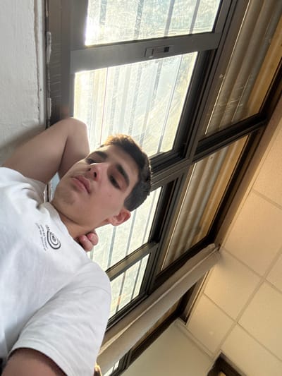 A young adult male, with dark short hair and light skin, is depicted in an indoor setting in Ness Ziona, Israel. He lies reclined, facing the camera with a neutral expression, his right arm bent with the hand positioned behind his head. He wears a white short-sleeved t-shirt featuring a dark circular graphic on the left chest. Behind him and to his right, a multi-paned window with dark framing allows bright sunlight to enter, revealing faint exterior details and partially obscuring light-colored vertical blinds or panels. A light-colored wall is visible to the left of the subject, and a suspended acoustic tile ceiling is visible in the upper right of the frame.