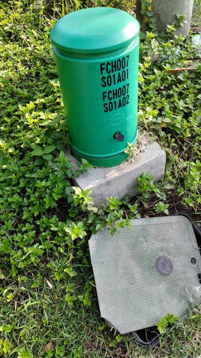 The image depicts an outdoor scene in Sidodadi, Indonesia, focusing on a green, cylindrical utility box situated on a concrete base amongst lush green vegetation. The utility box has black lettering that reads "FCH007 S01A01" and "FCH007 S01A02". Below this, a circular emblem with text is visible, which appears to say "Linknet". There is a small black lock mechanism on the side of the cylinder. To the right of the cylinder and slightly in front, a green, square utility box lid is resting on the ground, partially obscuring some of the vegetation. This lid has a textured surface and a circular emblem with writing. The surrounding environment is characterized by dense green ground cover, grass, and some taller green foliage. The lighting suggests it is daytime, with sunlight casting shadows. There are no people or animals visible in the image.