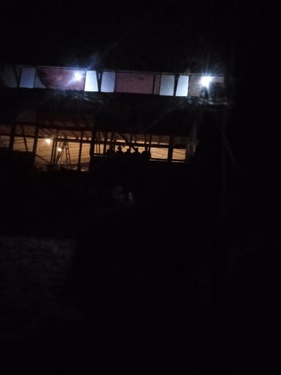 The image displays a structure in Al-Mawasi, Palestinian Territories, observed at night. The scene is predominantly dark, with internal illumination defining the building. The upper section of the structure presents multiple rectangular openings; some are covered with translucent reddish material, while others emit bright white light, creating visible glare. The lower level features a series of framed openings through which a warm, yellow-orange light emanates. Within this illuminated lower interior, the silhouettes of approximately five to six individuals are discernible, positioned towards the center-right. A ladder is visible leaning against an interior wall on the left side of the lower level. The immediate foreground and the surrounding environment are indistinct due to the pervasive darkness.