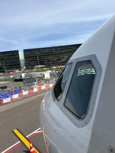 The image captures a detailed view of the nose and cockpit windows of a white or light gray commercial aircraft, positioned on an airport apron. The perspective is from an elevated point, looking forward and slightly down, with the aircraft's nose pointing towards the right. The cockpit windows, secured by visible rivets or screws, reflect elements of the sky and ground.
To the left of the aircraft, a large, modern building with a dark, gridded, reflective facade rises against a light blue sky, suggesting clear weather. Reflections of clouds can be seen on the building's surface.
Below and to the right of the aircraft, the asphalt of the airport apron is visible, marked with distinctive yellow, black, red, and white lines. An orange and white striped safety cone sits on a yellow and black striped ground marking in the lower right foreground.
Behind a chain-link fence on the apron, several service vehicles are present. Two prominent white trucks, clearly branded with the text "eat.gourmet," are visible, indicating catering services. Other trucks, trailers, and stacks of colorful containers (red, blue, white) are also arranged in the service area.
The scene depicts a daytime setting at an airport, likely during ground operations such as servicing or preparation for a flight. The overall impression is one of an active, organized airport environment in Kloten, Switzerland.