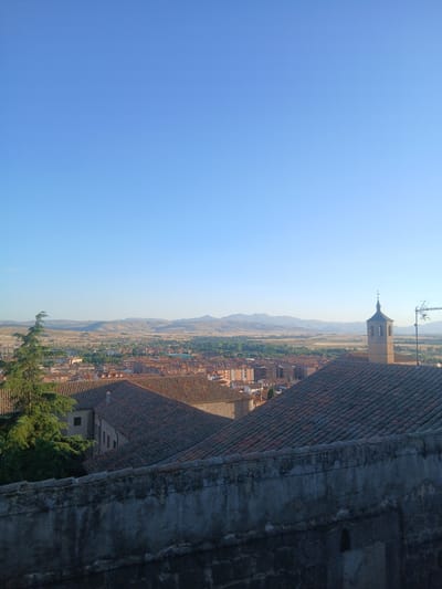 The image presents a panoramic view of the city of Ávila, Spain, likely taken from an elevated vantage point. The foreground is dominated by a weathered stone wall, with a visible texture of bricks and a rough, greyish finish. Beyond this wall, the roofs of numerous buildings stretch out, characterized by their terracotta tiles and varied architectural styles, suggesting an old European city.
A prominent feature in the mid-ground is a church bell tower, with a distinctive steeple, topped with a cross. Its stone construction and architectural details suggest historical significance. Adjacent to the bell tower, an antenna is visible on a rooftop, indicating modern infrastructure within the historic cityscape.
The mid-ground also reveals a lush green tree with a prominent trunk and dense foliage, partially obscuring some of the buildings. This tree adds a natural element to the urban landscape.
The background of the image shows a sprawling urban area with buildings of varying heights and colors. Beyond the city, rolling hills and distant mountains form a natural horizon. The sky above is a clear, bright blue, suggesting a sunny day with excellent visibility. The overall impression is one of a historic city nestled within a scenic natural environment.