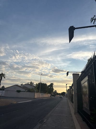 This is an eye-level, outdoor, medium shot of a street in Pia, France at dusk. The sky is a gradient of blue with scattered white clouds. On the left side of the road are houses and palm trees. The road is asphalt with white lane markings. A sidewalk runs along the right side of the road, bordered by a black metal fence and a stone wall. Two cars are visible on the road, one white and one dark colored. Traffic lights are visible suspended over the road. The overall lighting suggests it is late afternoon or early evening, with the sun setting.