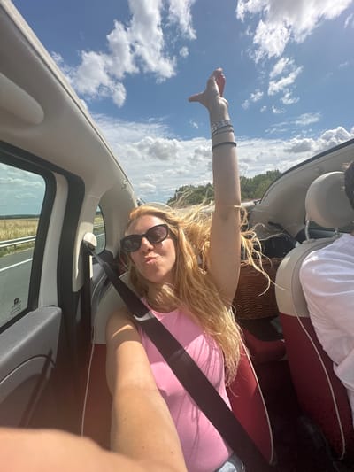A woman with long blonde hair and sunglasses is leaning forward in the passenger seat of a convertible, her right arm raised with her hand pointing upwards. She is wearing a pink tank top and multiple bracelets on her raised arm. Her left arm is also visible, extended forward, likely holding a phone for a selfie. The car's interior is visible, with a dark grey dashboard and red and white seats. A wicker basket is seen in the back seat. The car appears to be on a road with a field and trees visible in the background under a bright, cloudy blue sky. The scene suggests a journey or road trip. There is no specific indication of the location being Alaincourt, France.