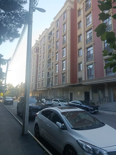 The image depicts a street scene in Sumgayit, Azerbaijan, in front of a modern, multi-story building. The building has a facade of red brick and beige stone accents, with numerous windows and balconies. A row of parked cars lines the street, including a white sedan in the foreground, a black pickup truck, and other vehicles further down the road. Trees and utility poles are visible along the sidewalk. The lighting suggests it is daytime, with the sun casting shadows and creating highlights. The environment appears to be an urban residential or commercial area. No people are prominently visible in the frame.